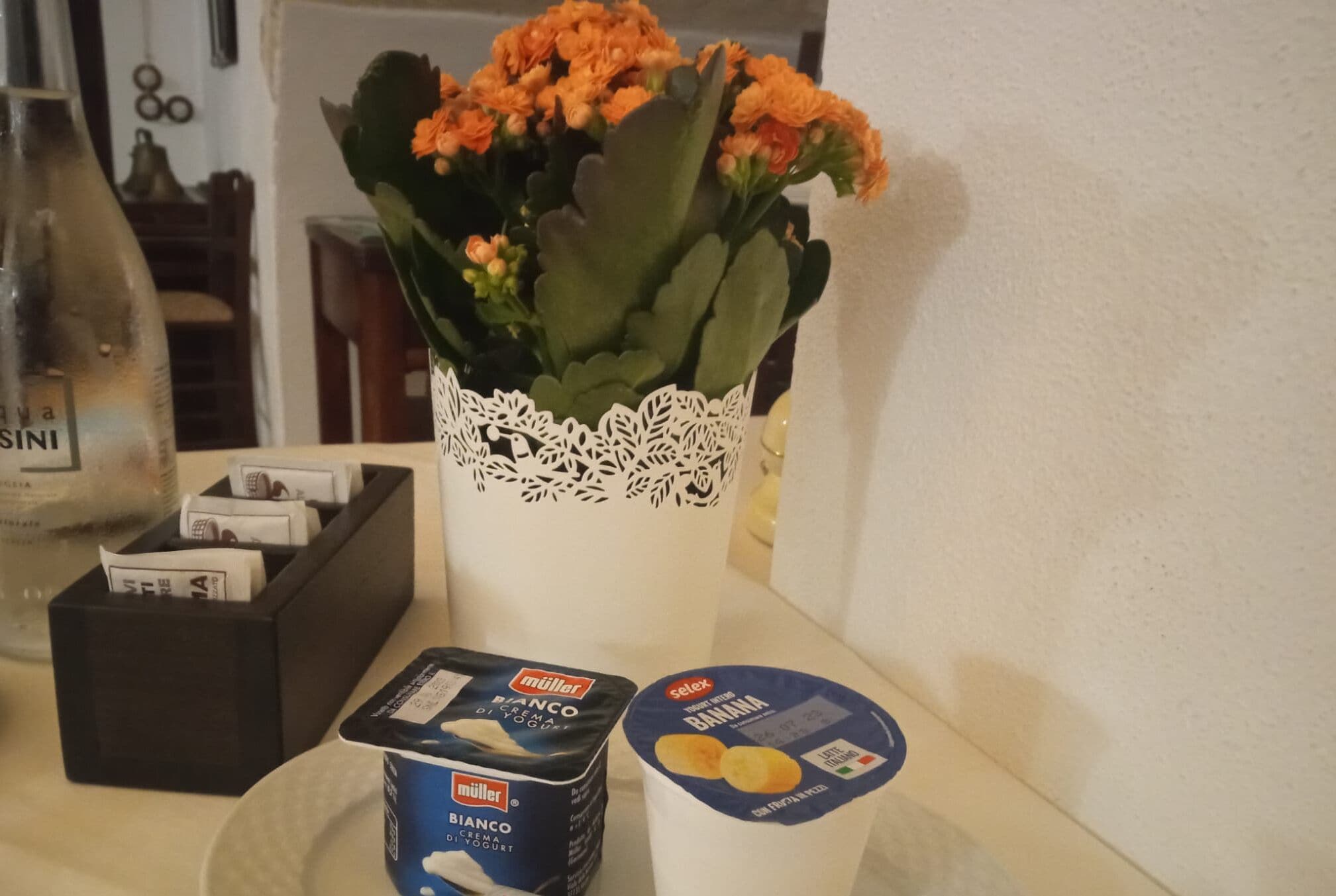 A table with a white lace vase containing orange flowers, a black box with money, and two containers of Müller yogurt.