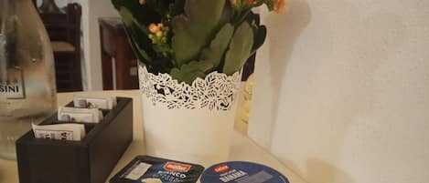 Una mesa con un florero blanco de encaje con flores naranjas, una caja negra con dinero y dos recipientes de yogur Müller.