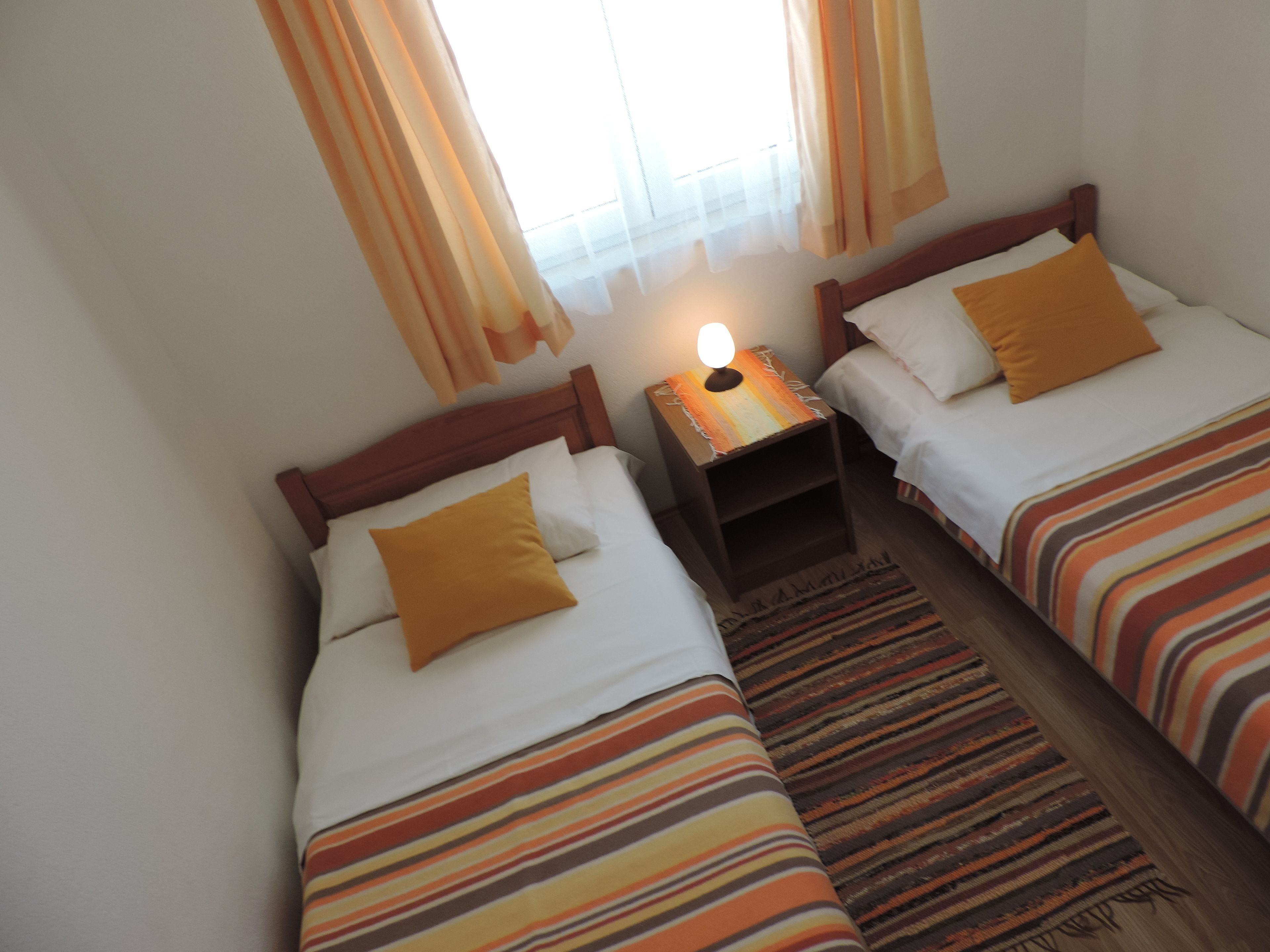 A hotel room with two single beds, each with a wooden headboard, white bedding, and a striped orange and brown bedspread. There is a nightstand with a lamp between the beds, and a window with sheer curtains.