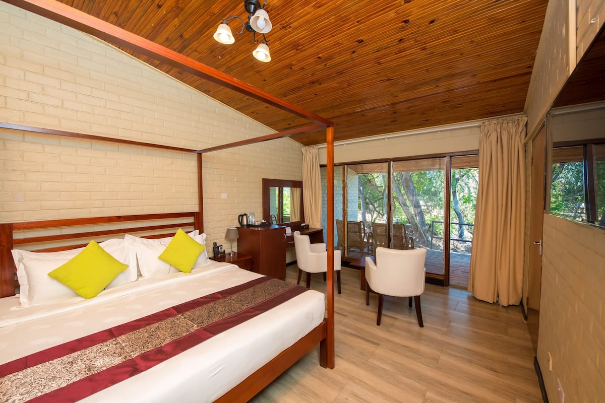 A four-poster bed with a wooden headboard, a desk with a chair, and a view of the outdoors through large windows.