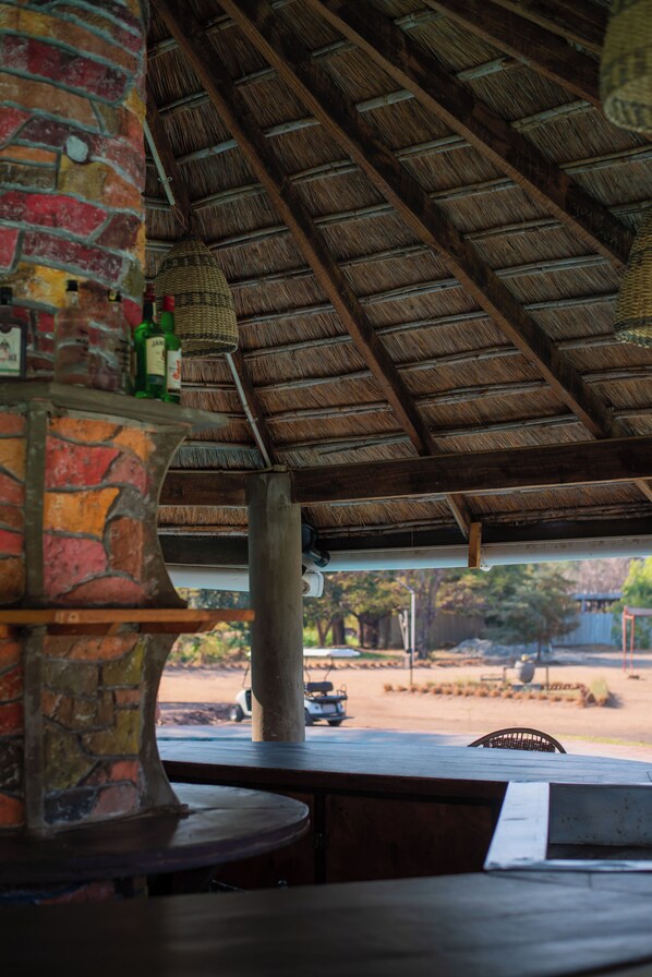A rustic outdoor seating area with a thatched roof, brick walls, and a view of a dirt path and trees.
