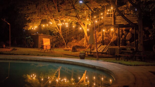 A nighttime outdoor setting with a swimming pool, wooden deck, lounge chairs, and string lights.