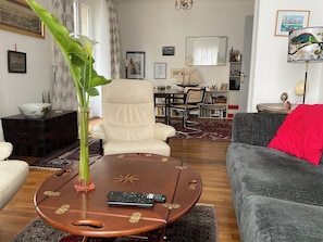 A living room with a round wooden coffee table, a beige armchair, a grey sofa, a large green plant, and a bookshelf with various items.