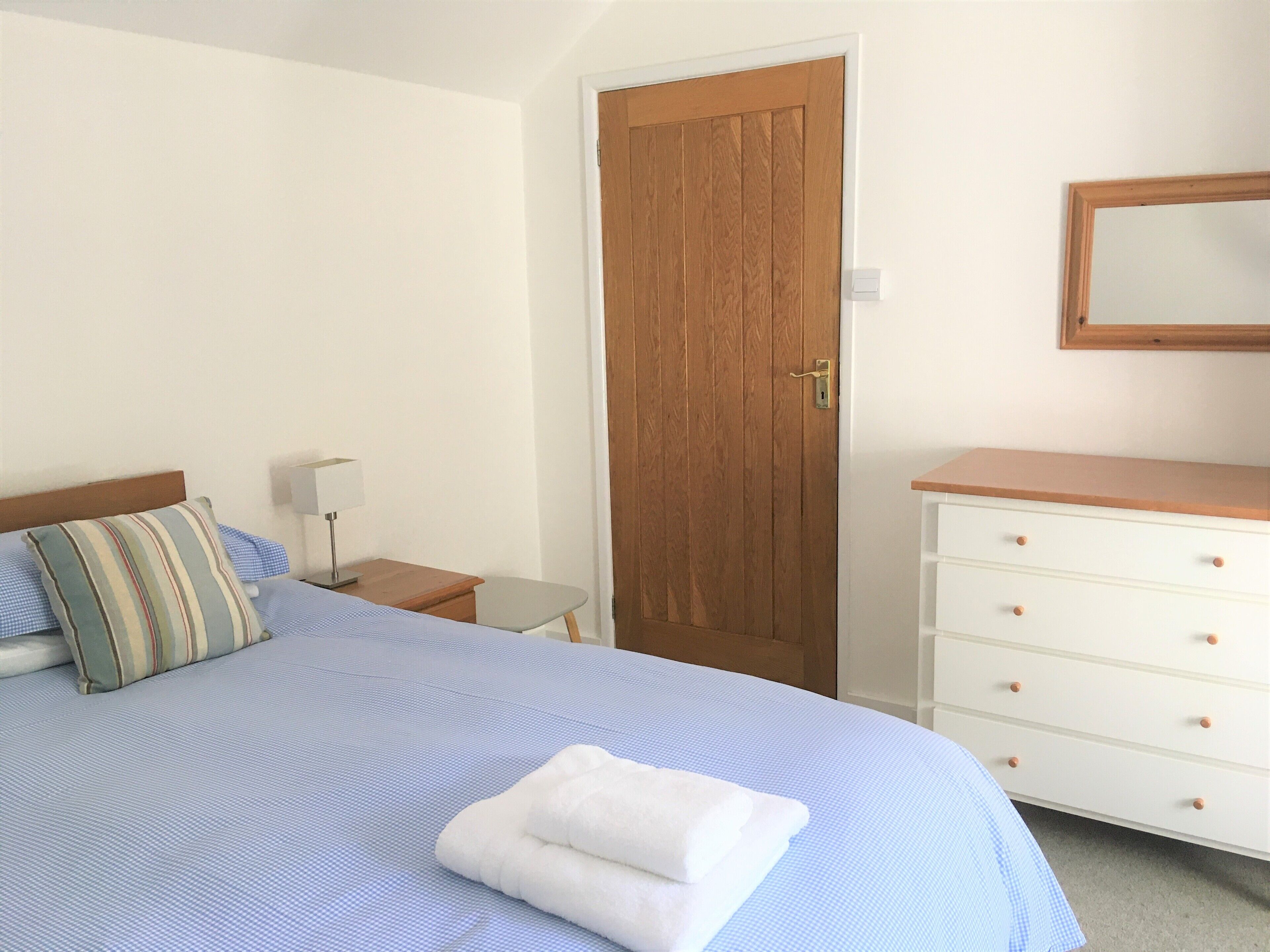 Un dormitorio con cama, una puerta de madera, una mesita de noche, un aparador y un espejo.