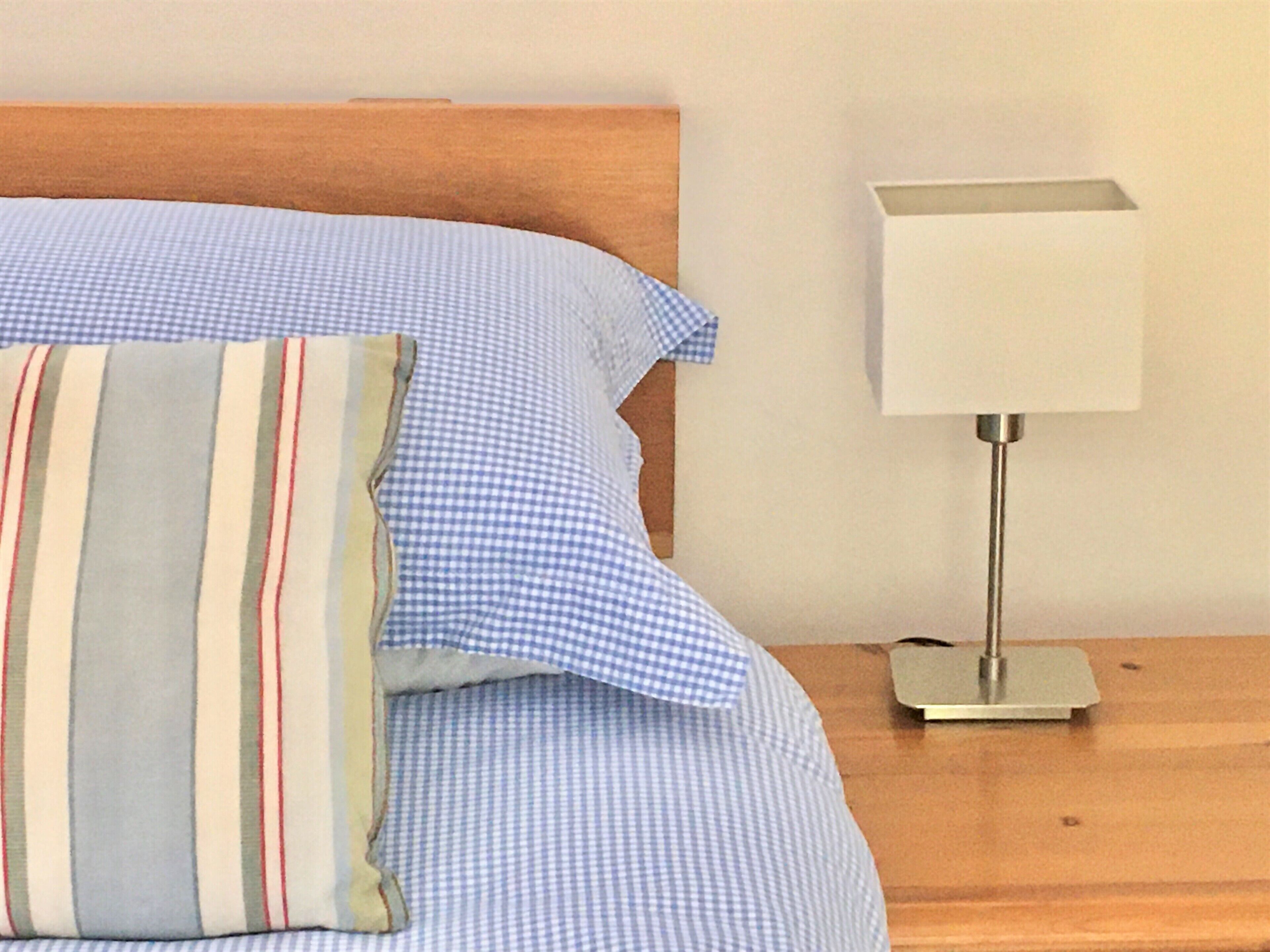 A neatly made bed with blue checkered sheets and striped pillows, a bedside table with a lamp, and a wooden headboard.