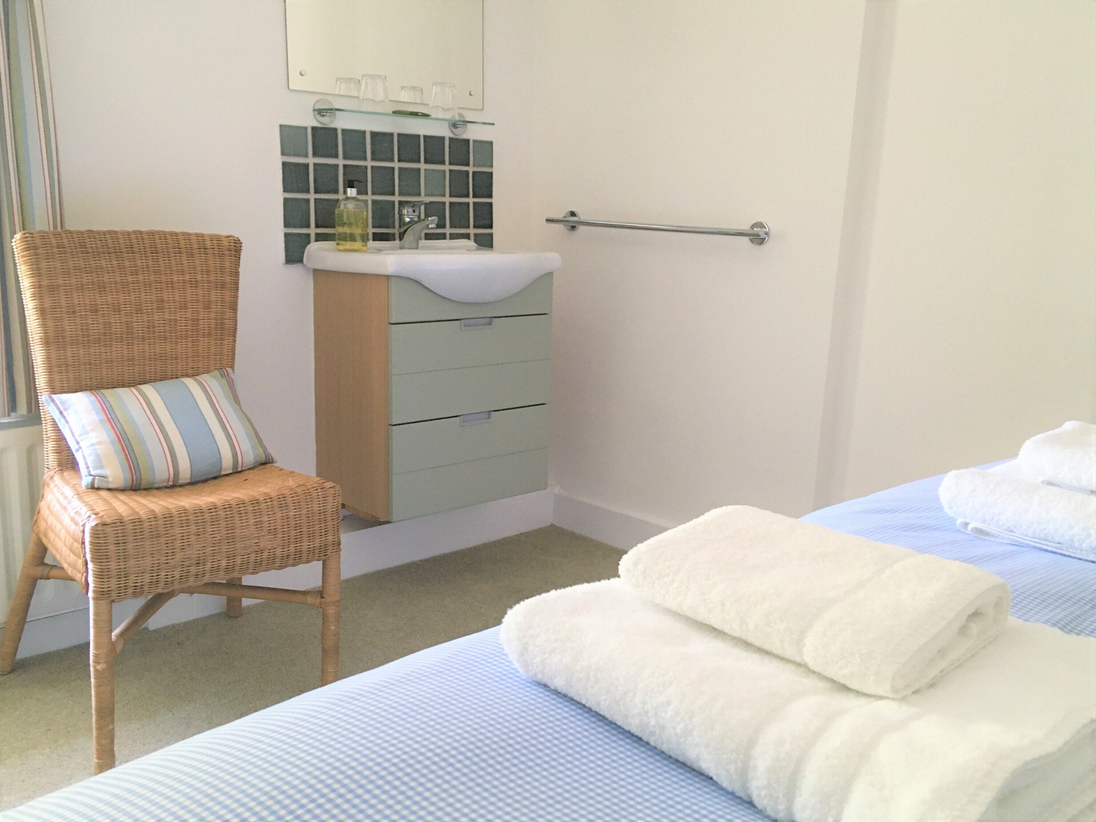 A bedroom with a bed, a chair, a bedside table, a mirror, and a bathroom with a sink and a towel rack.