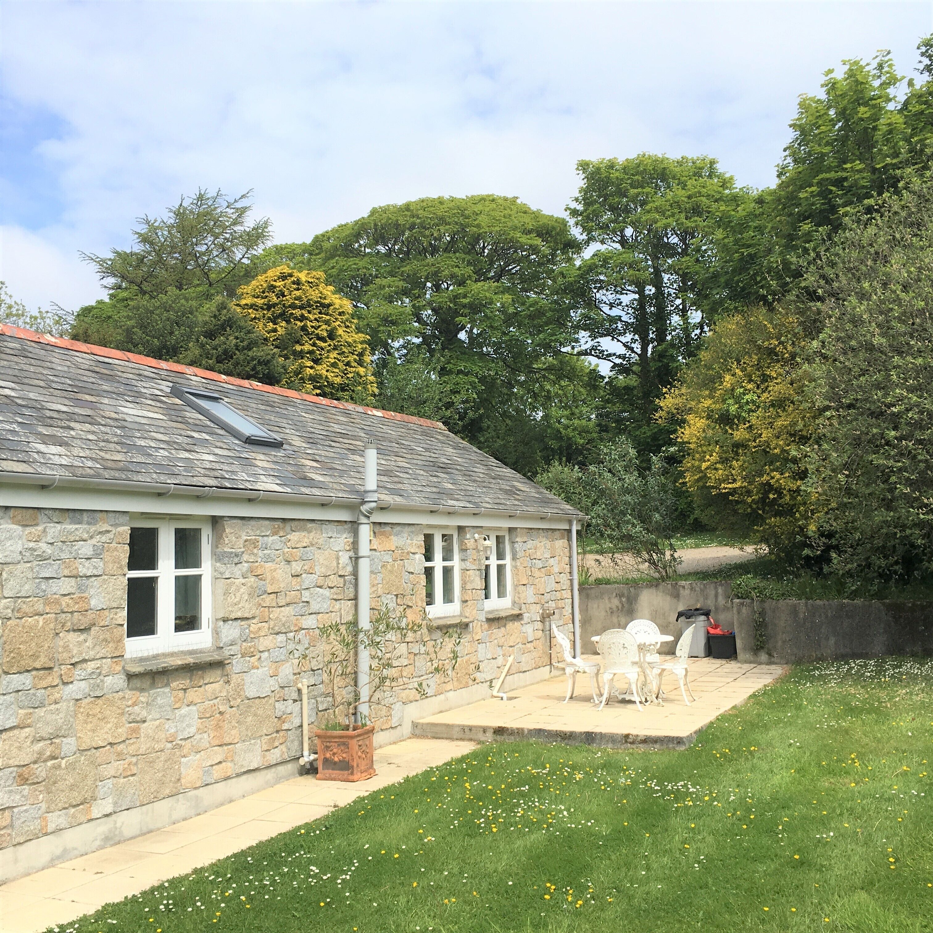 A stone house with a tiled roof, a small patio with white chairs, and a garden with grass and flowers.