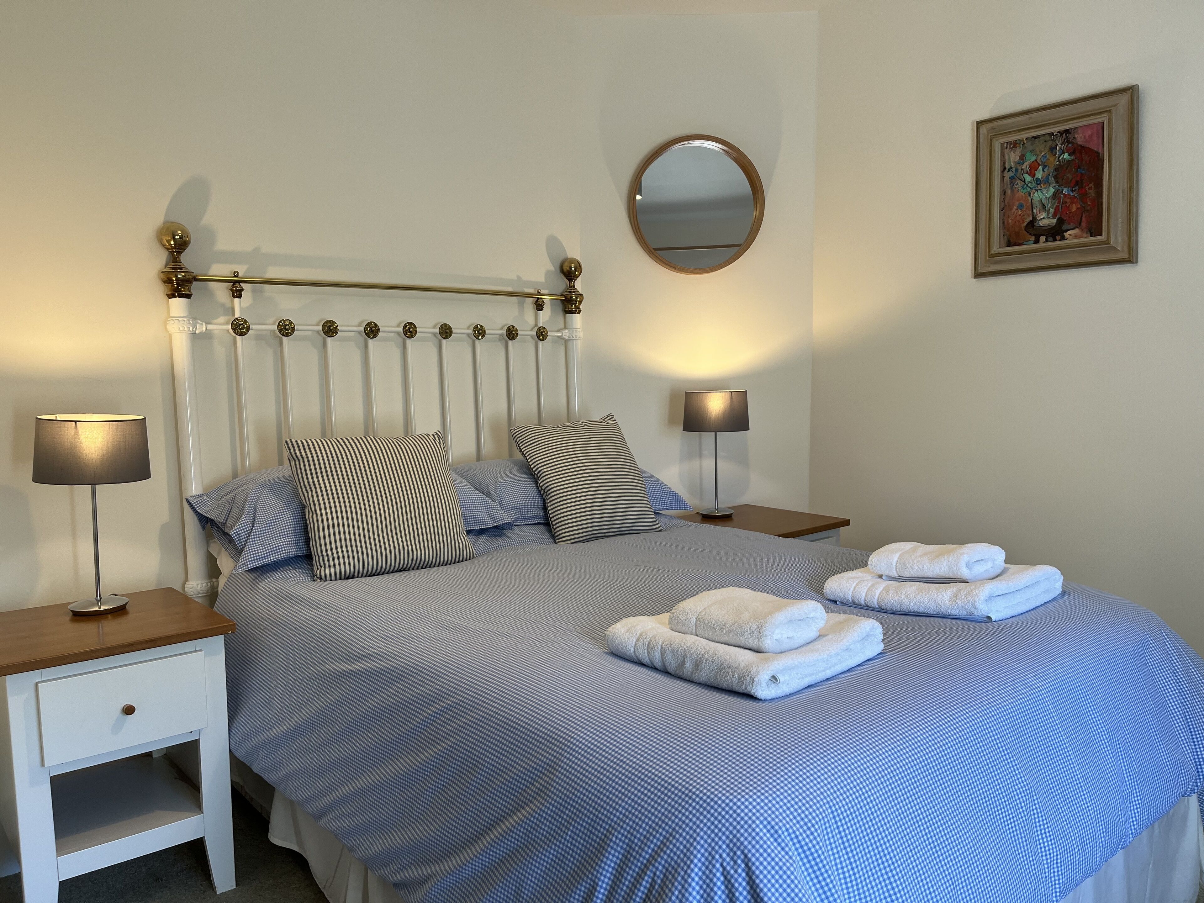 A neatly made bed with blue bedding, two bedside tables with lamps, a round mirror, and a framed artwork on the wall.