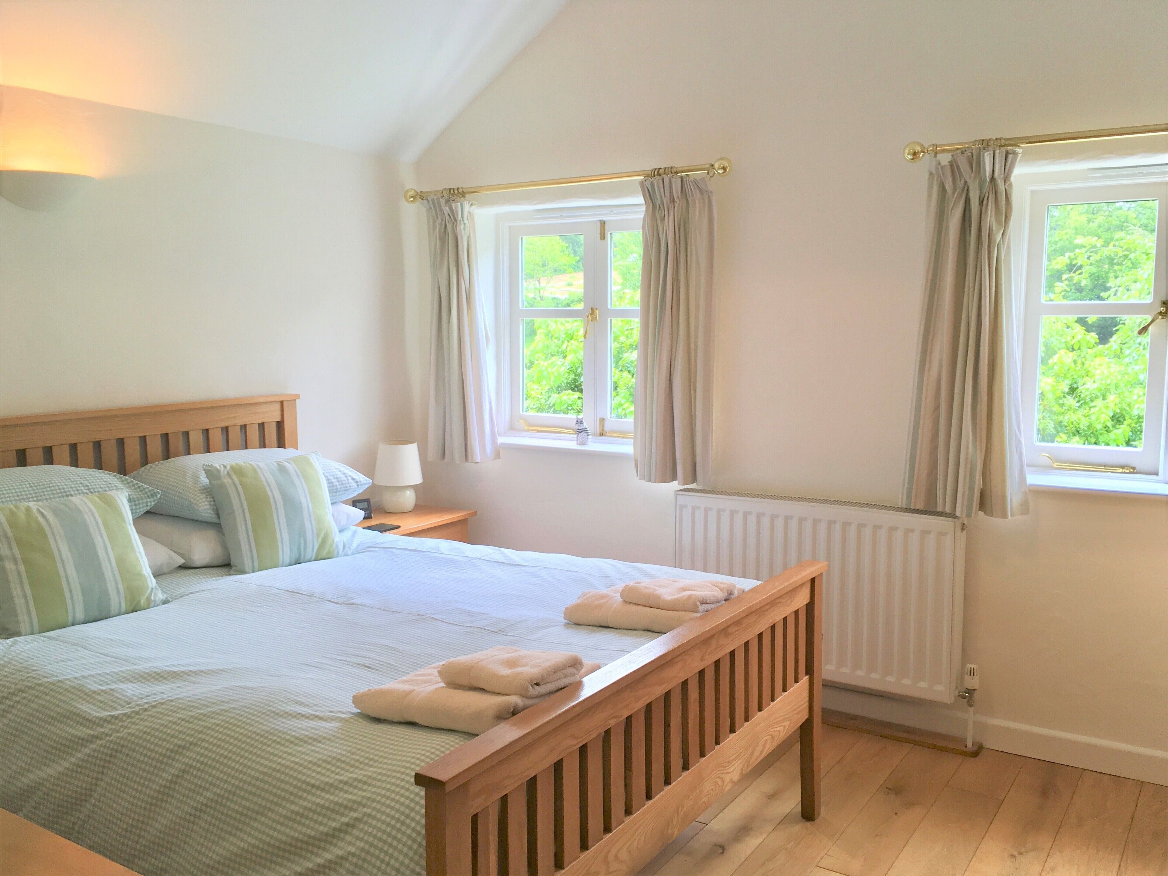 A bedroom with a wooden bed, a nightstand with a lamp, two windows with curtains, and a view of greenery outside.