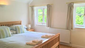 A bedroom with a wooden bed, a nightstand with a lamp, two windows with curtains, and a view of greenery outside.