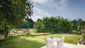 A patio with a white table and chairs, surrounded by a lush garden and a wooden fence. - Little Pengelly Farm (Hayle)