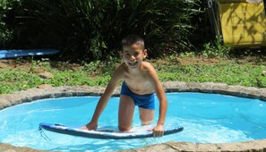 Un enfant se trouve dans une petite piscine ronde, hors sol, avec une planche gonflable bleue, entouré de verdure et avec une structure jaune en arrière-plan.