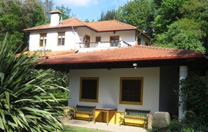 Une maison à deux étages, avec un toit recouvert de tuiles rouges, un balcon et deux fenêtres aux encadrements jaunes.