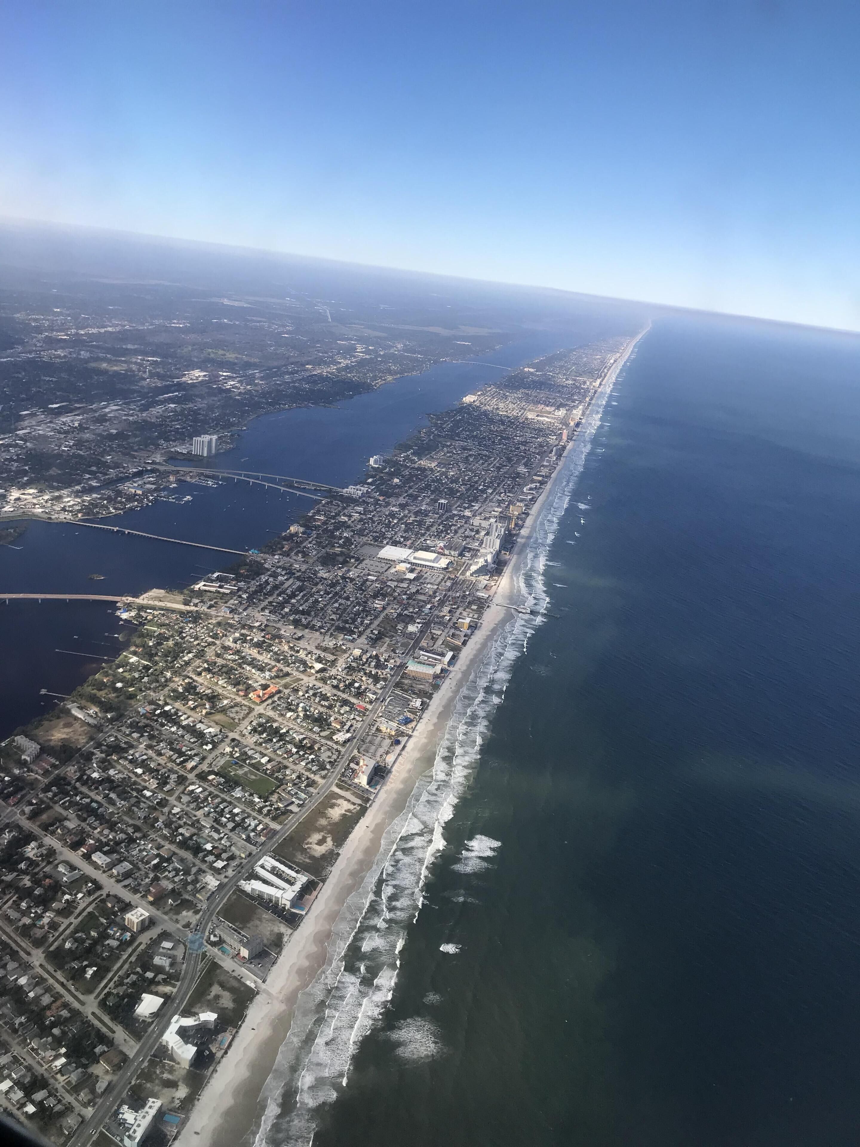 Vista aérea de una ciudad costera con una extensa playa, una zona urbana densa y un puente que une dos porciones de tierra.