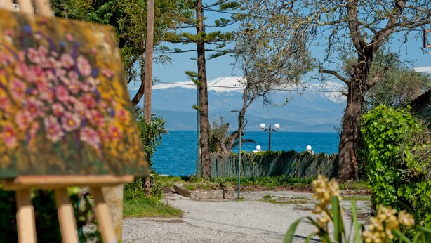Ein malerischer Blick auf eine Küstenlandschaft mit einem Gemälde auf einem Malkasten, Bäumen und einem klaren Blick auf das Meer und die Berge im Hintergrund.