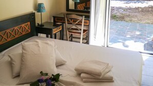 Ein ordentlich bezogenes Bett mit Kissen und einem gefalteten Handtuch, ein Schreibtisch mit Stuhl und ein großes Fenster mit Blick ins Freie.