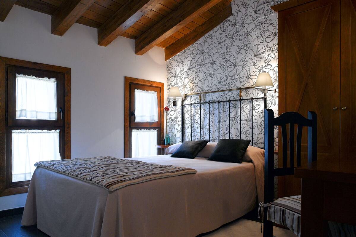 Un dormitorio con cama, cabecera de madera, dos ventanas con cortinas, una mesita de noche y un armario de madera.