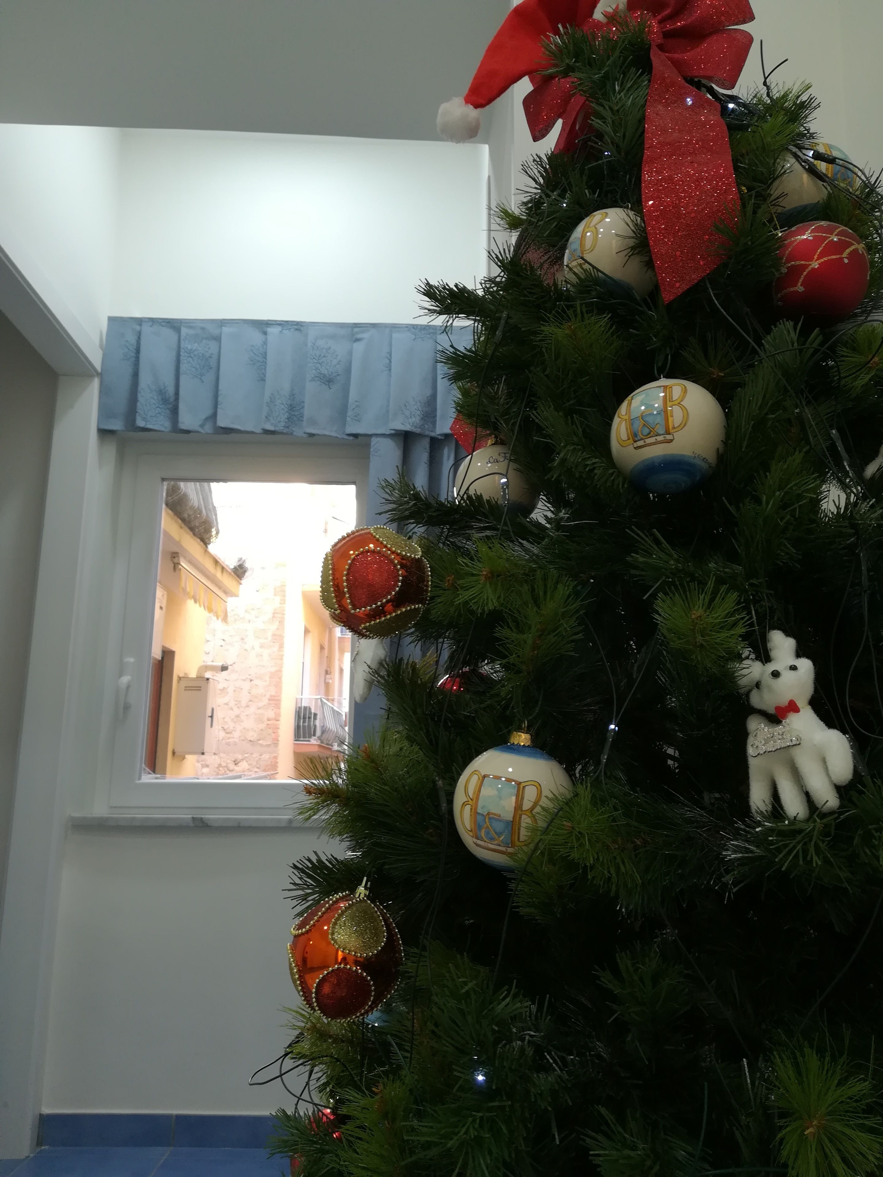 Ein Weihnachtsbaum mit Dekorationen und einem Teddy, platziert in der Nähe eines Fensters mit blauer Vorhang.