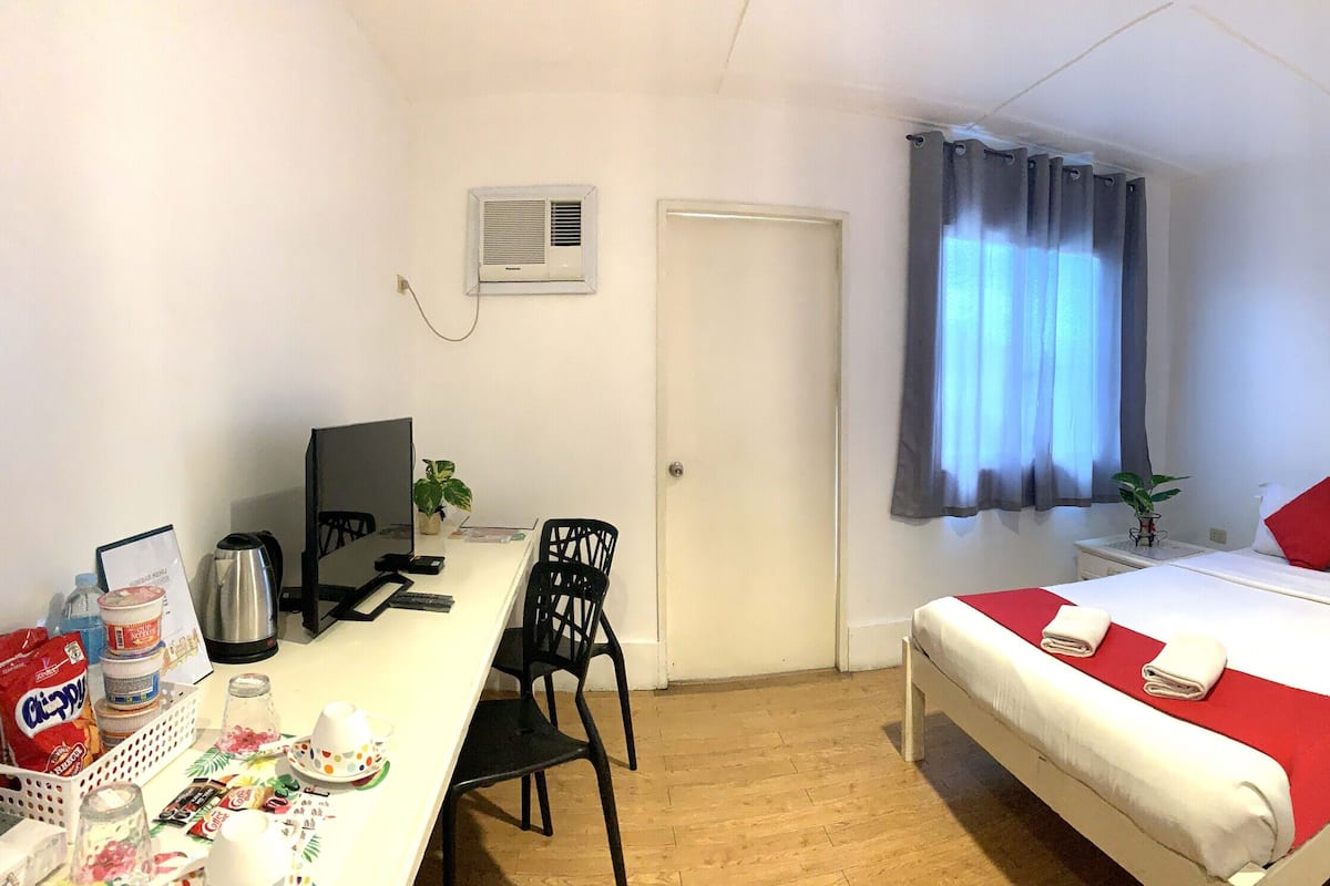 Une chambre d’hôtel comprenant un lit, un bureau avec un ordinateur, une chaise, une fenêtre avec des rideaux, un tableau au mur, ainsi qu’une salle de bain avec une douche et des toilettes.