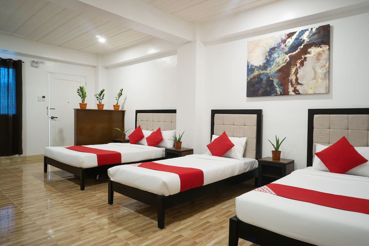 Une chambre d’hôtel avec trois lits, chacun recouvert d’une courtepointe rouge et blanche assortie aux coussins. On trouve des plantes en pot sur les tables de chevet et un grand tableau abstrait au mur.