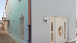 A building with a light blue exterior, a white door with a glass panel, and a sign with a logo and contact information. - Le mille e una notte (Comacchio)