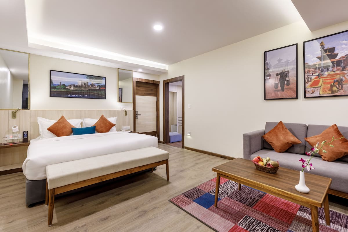 A modern hotel room with a large bed, a sofa, a small table with a vase and fruit basket, and two framed pictures on the wall.
