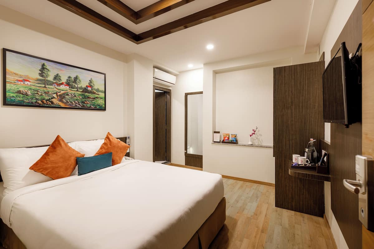 A hotel room with a bed, pillows, a painting, a wooden ceiling, a door, and a shelf with items.