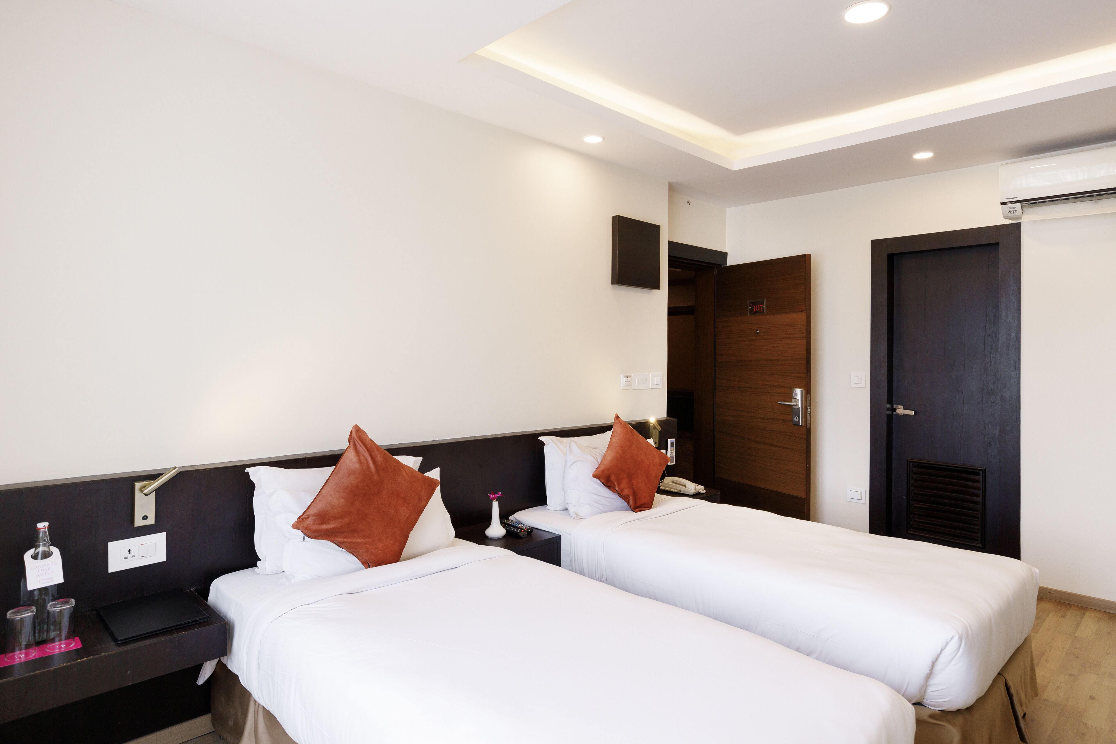 A hotel room with two single beds, a bedside table with a lamp, a small table with a bottle and glasses, and a wall-mounted air conditioning unit.