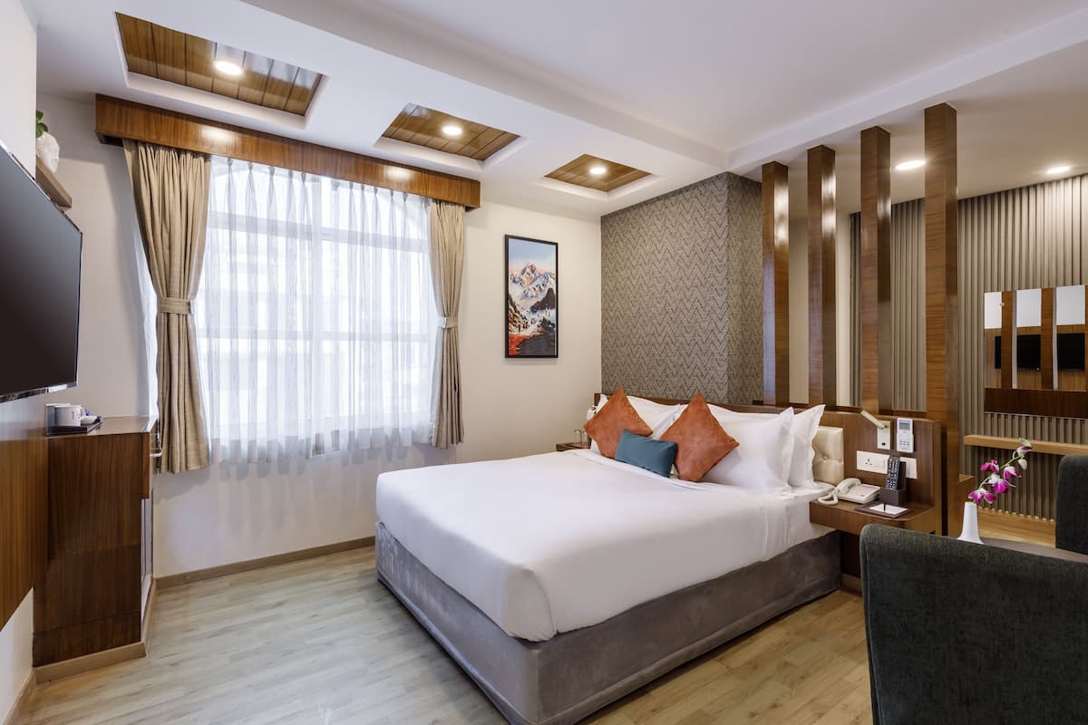 A hotel room with a large bed, a desk with a flat-screen TV, a bedside table with a phone, and a window with curtains.