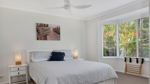 A bedroom with a bed, nightstands, a ceiling fan, a window with blinds, and a beach-themed artwork on the wall. - Beautiful , stylish Mudjimba Beach House (Mudjimba)