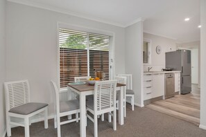 A compact kitchen with white cabinetry, a stainless steel refrigerator, and a dining area with white chairs and a wooden table. - Beautiful , stylish Mudjimba Beach House (Mudjimba)