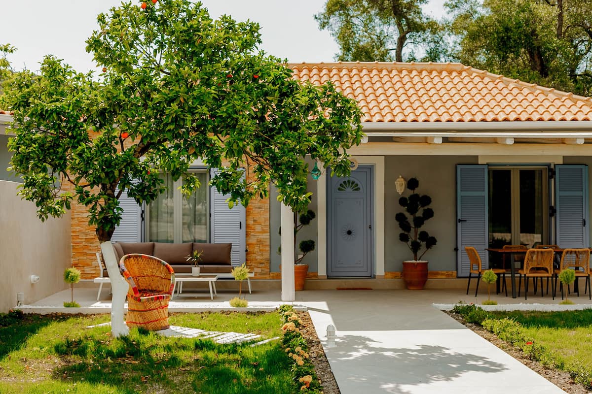 Una casa con techo de tejas, un jardín con un árbol, mobiliario exterior y una puerta con contraventanas.