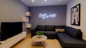 Un salon moderne avec un téléviseur à écran plat, un canapé gris, une table basse ronde ornée d’une plante et un néon mural.