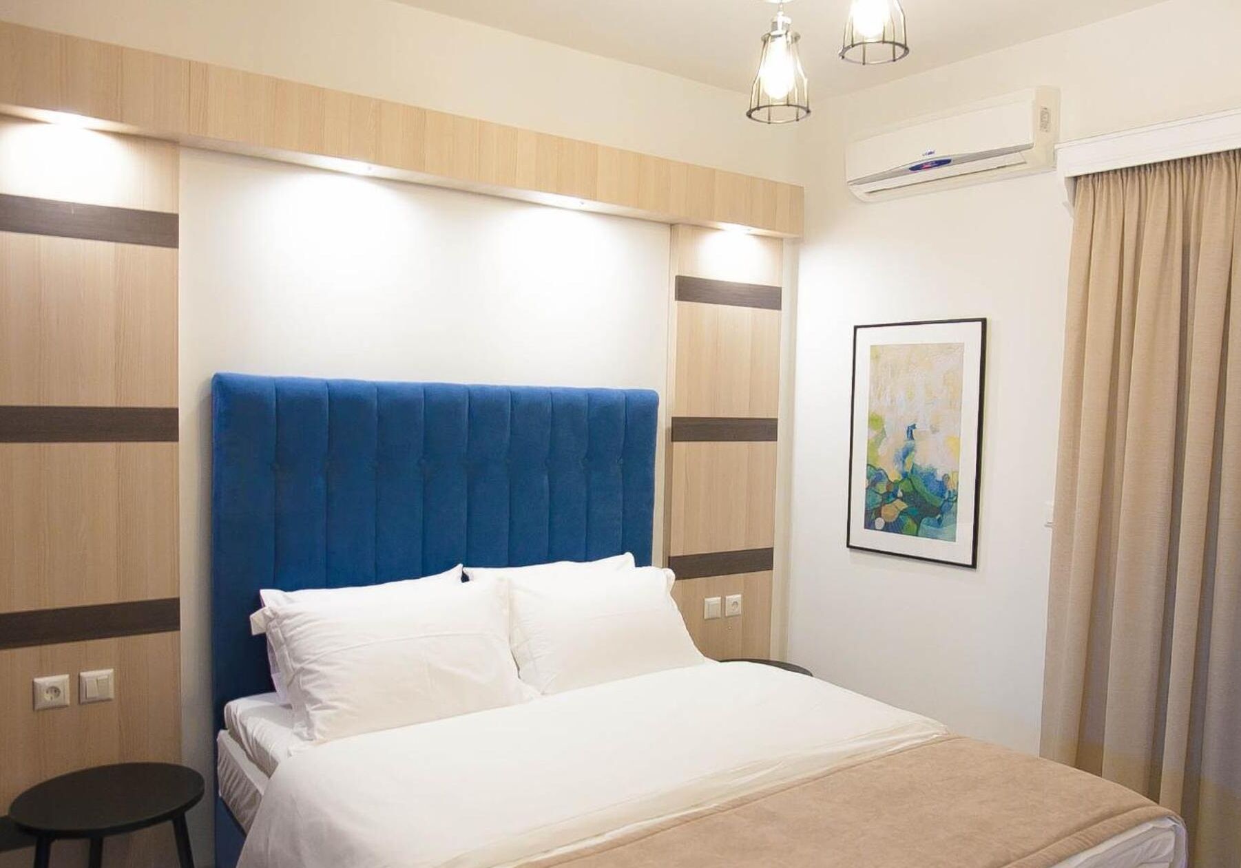 A hotel room with a bed, a bedside table, a wall-mounted air conditioner, and a framed abstract painting.