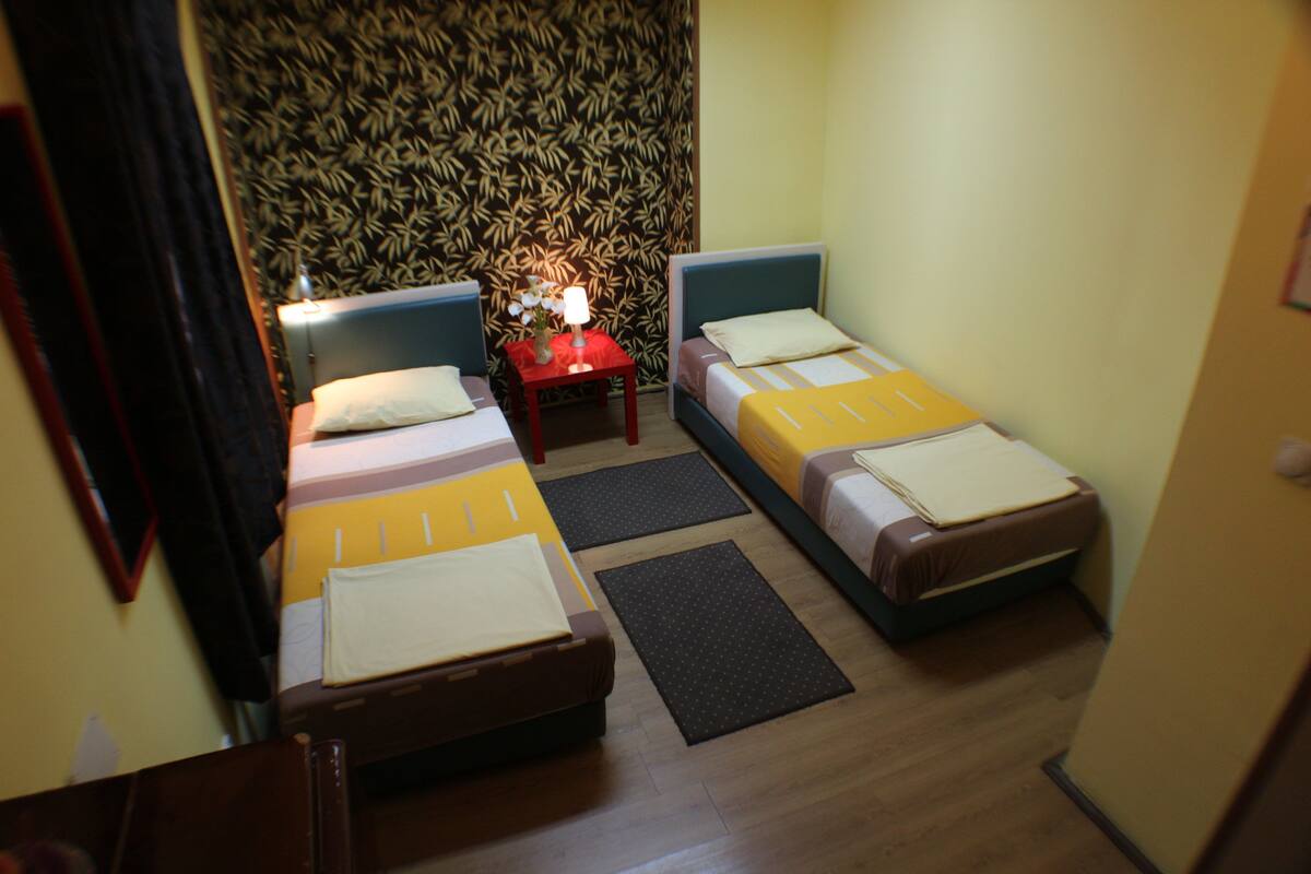 A hotel room with two single beds, a small table with a lamp, and a patterned wallpaper.