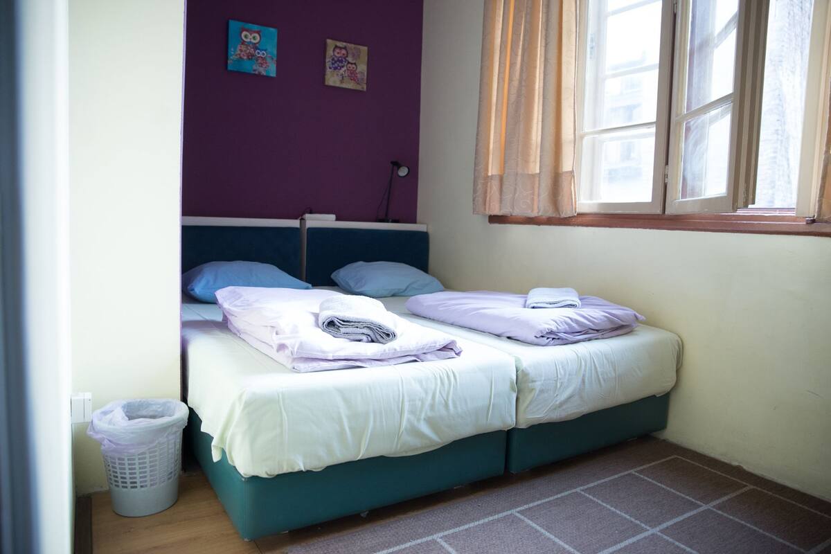 A small, neatly made bed with two pillows and a folded blanket, a laundry basket, and a window with curtains.
