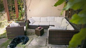 Un patio con mobiliario de mimbre, incluyendo un sofá y dos sillones, y una mesita. Un perro negro está acostado en el suelo.