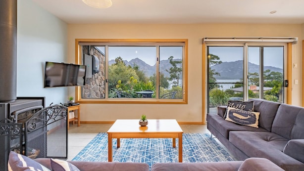 A living room with a sofa, a coffee table, a TV mounted on the wall, and large windows offering a view of mountains and greenery. - Greystone (FS5) - Freycinet Holiday Houses (Coles Bay)