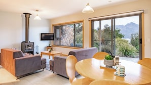 A modern living room with a stone fireplace, comfortable seating, a dining area, and large windows offering a mountain view. - Greystone (FS5) - Freycinet Holiday Houses (Coles Bay)