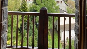 Ein Fenster mit Steinrahmen und Holzbalkon, das einen Blick auf eine gebirgige Landschaft mit Gebäuden und Grünflächen bietet.