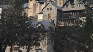 Ein historisches Steinhaus mit einem markanten Turm, gelegen in einer gebirgigen Landschaft mit einer großen Felsformation im Hintergrund.