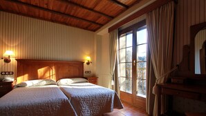 Ein Zimmer mit zwei Betten, Holzbalkendecke, einem großen Fenster mit Vorhängen und einem Holztisch.