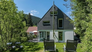 A modern two-story house with a sloped roof, large windows, and a visible chimney. There are two lawn chairs in the foreground. - Luxus Ferienhaus Schwarzwald Dieboldsberg (Alpirsbach)