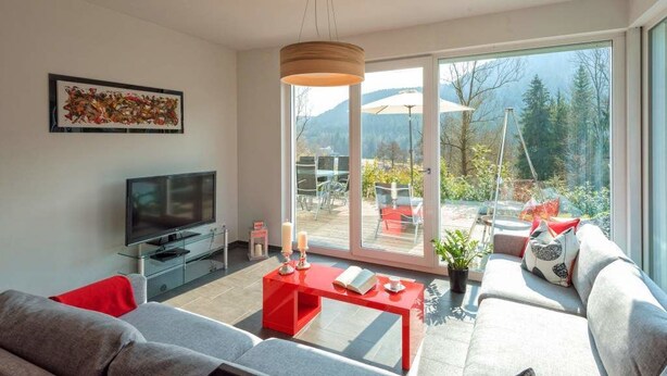 A modern living room with a TV, red coffee table, and a sofa. It features large windows with a view of a garden and mountains. - Luxus Ferienhaus Schwarzwald Dieboldsberg (Alpirsbach)