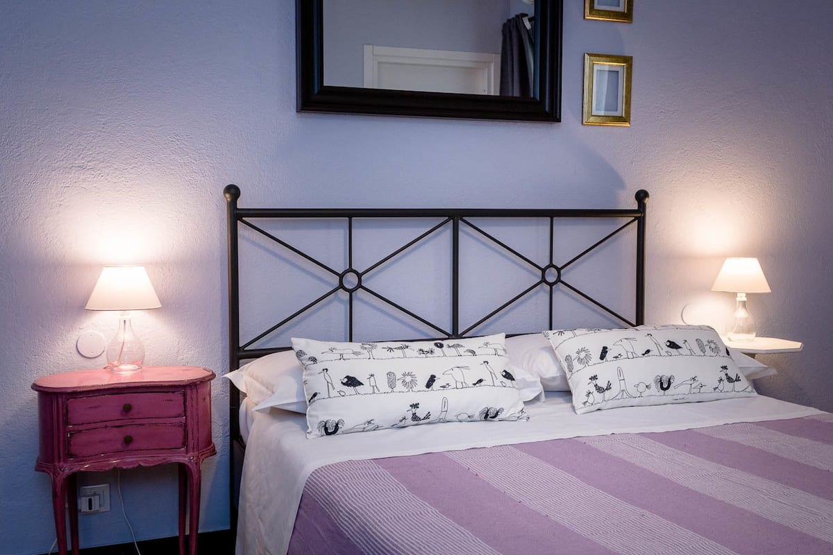 Una cama con cabecera metálica, dos mesitas de noche con lámparas y almohadas estampadas.