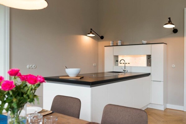 Una cocina moderna con armarios blancos, encimera negra y un lavamanos empotrado. Hay una mesa de comedor con sillas y un florero con flores.