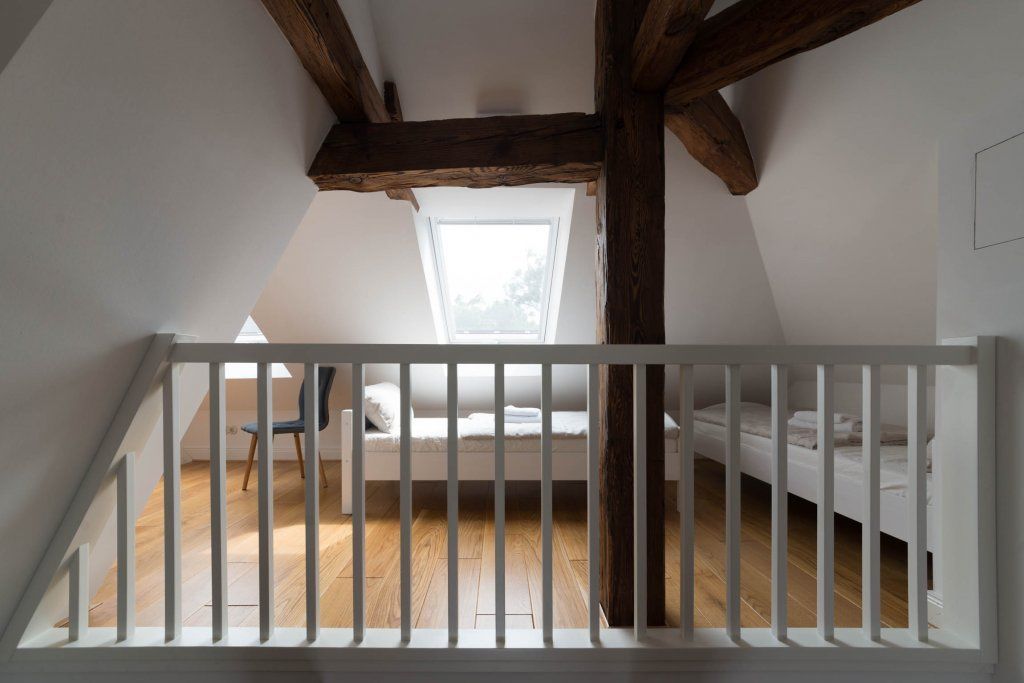 A dormitory room with a sloped ceiling, wooden beams, a skylight, and a white railing.