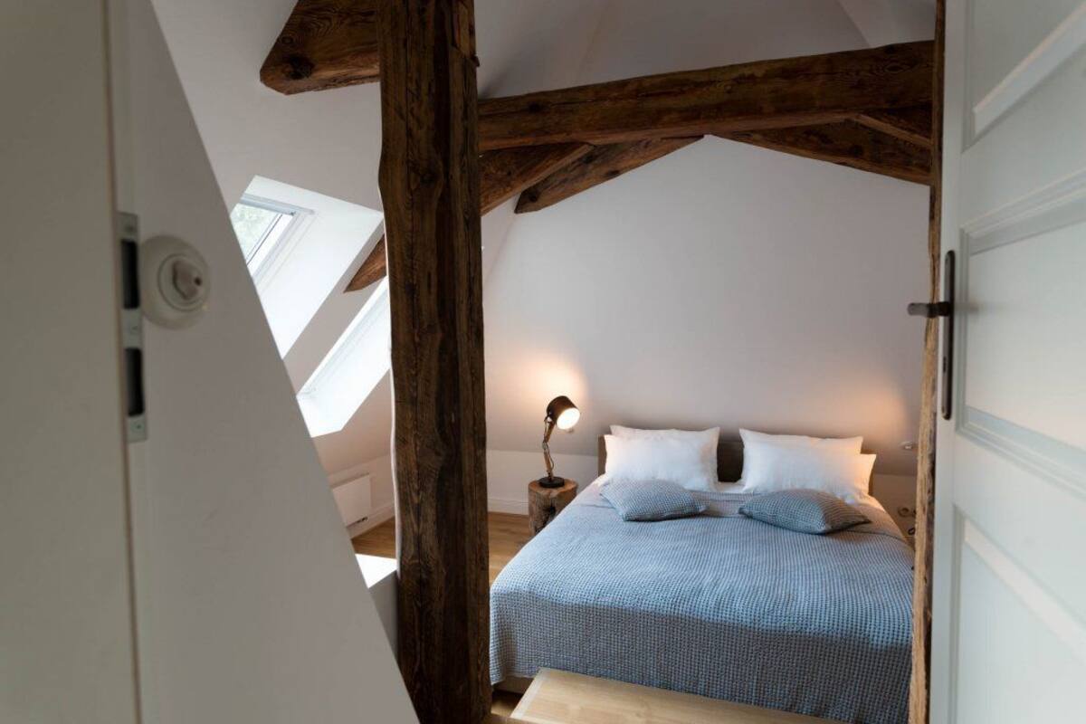 Une chambre avec un plafond en pente, des poutres apparentes, un lit recouvert d’une couverture à carreaux bleus, des oreillers blancs et une lampe de chevet.
