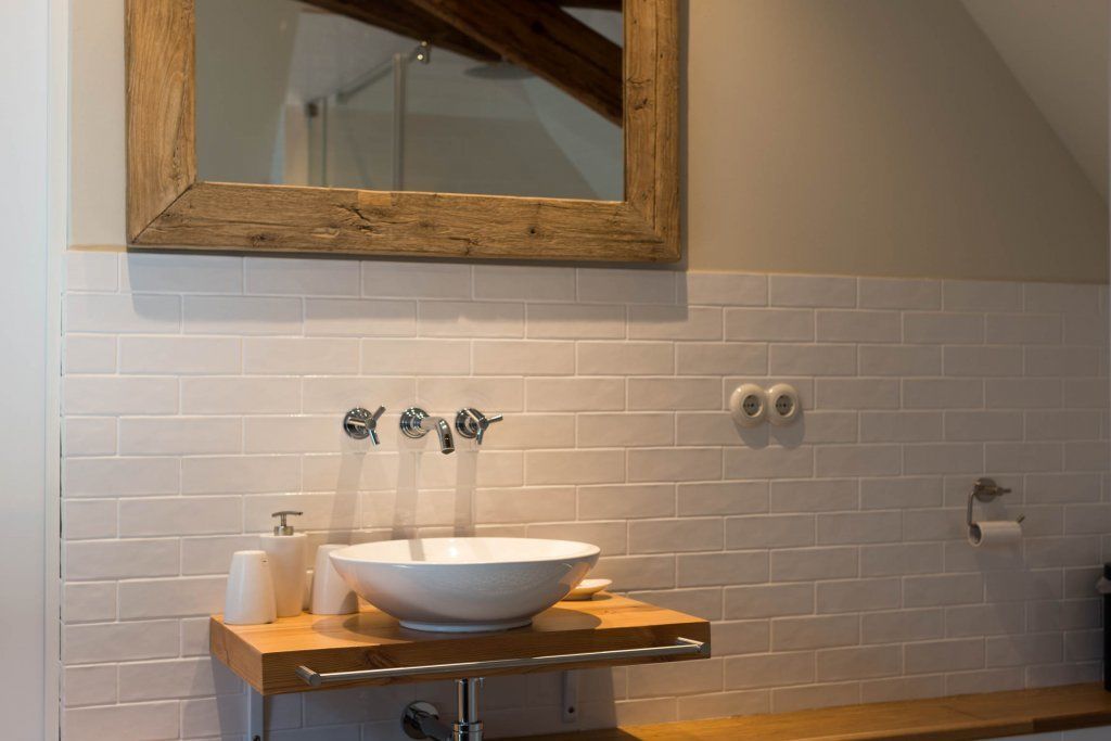 A bathroom with a white tiled wall, a wooden-framed mirror, a white sink with a wooden countertop, and a towel dispenser.