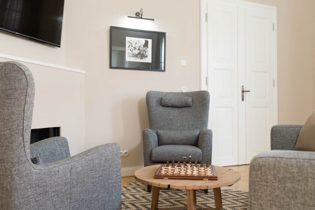 Un salón con sillones tapizados de gris, una mesa de centro de madera con un tablero de ajedrez, un cuadro en la pared y una puerta cerrada.